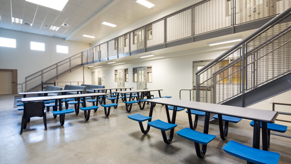 Empty institutional common area with cafeteria tables, blue seats, metal staircases, and a second-floor walkway with cell doors