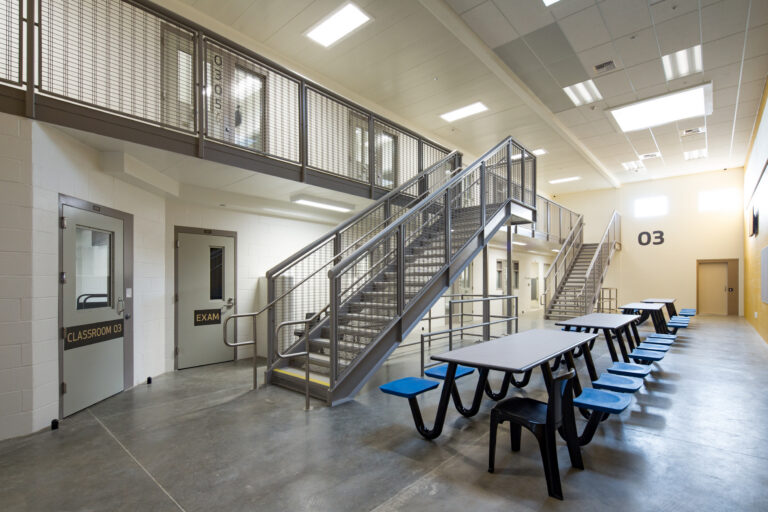 Interior of a modern detention facility with metal stairs, upper cell walkway, and cafeteria tables near doors labeled Classroom 03 and Exam