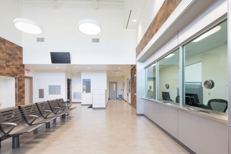 Modern waiting area with metal benches, high ceiling, and a glass reception window in a bright office lobby