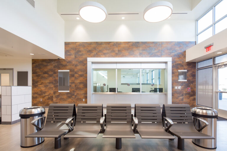Modern waiting area with rows of chairs, stainless steel trash cans, and a reception window beneath tall ceiling lights.
