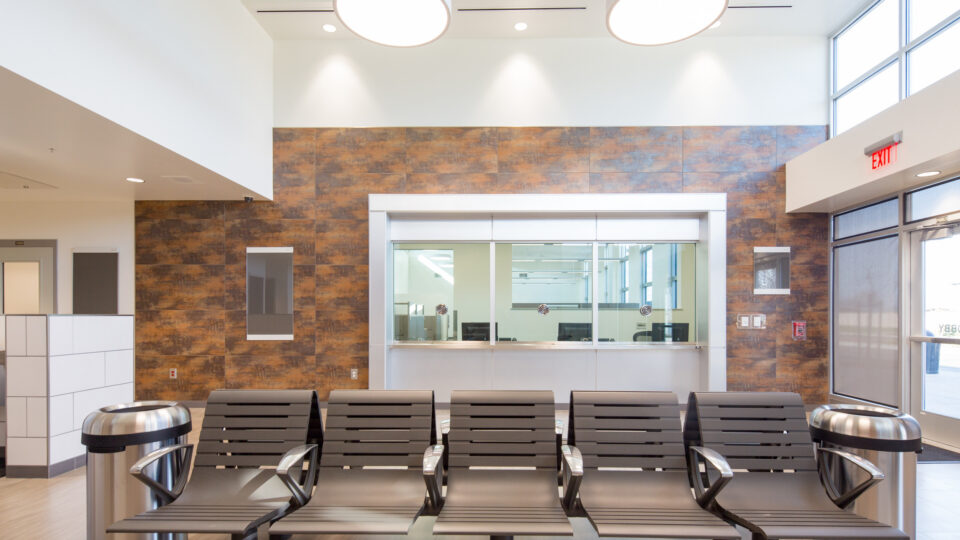 Modern waiting area with rows of chairs, stainless steel trash cans, and a reception window beneath tall ceiling lights.