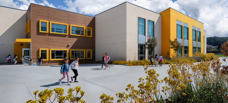 Children walking outside a modern school building with yellow accents, large windows, and landscaped grounds under a blue sky.