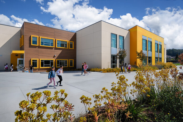 Children walking outside a modern school building with yellow accents, large windows, and landscaped grounds under a blue sky.