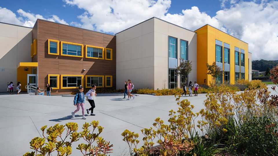 Children walking outside a modern school building with yellow accents, large windows, and landscaped grounds under a blue sky.