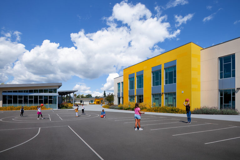 Children playing with basketballs on a school playground beside a modern yellow-and-white building under a bright blue sky.