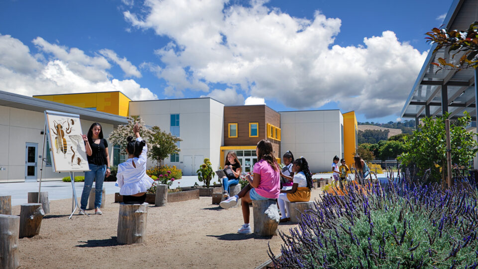 Teacher leads children in an outdoor lesson by a bee diagram in a school garden courtyard.
