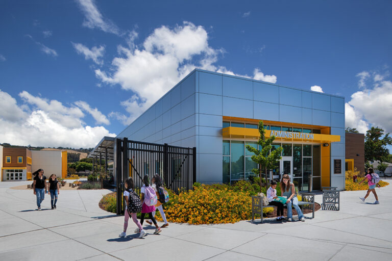 Modern school administration building with yellow accents, students walking outside, and a teacher reading with a child on a bench.