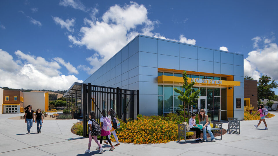 Modern school administration building with yellow accents, students walking outside, and a teacher reading with a child on a bench.