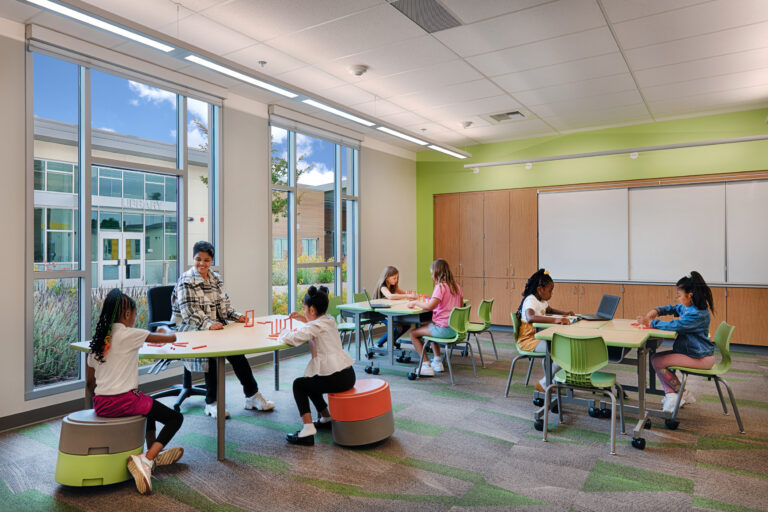 Teacher and young children working at tables in a bright classroom with large windows and a library visible outside