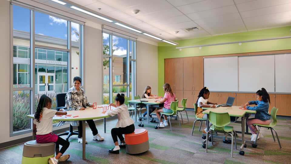 Teacher and young children working at tables in a bright classroom with large windows and a library visible outside