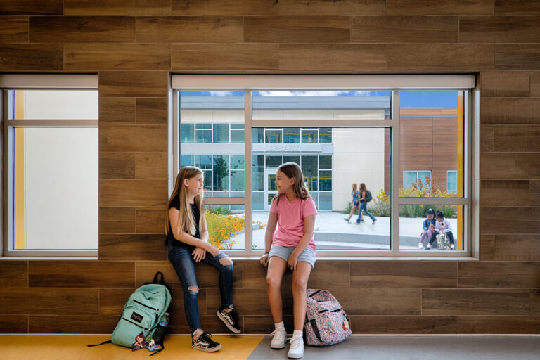 Two students sit on an indoor window ledge with backpacks, talking as a school library courtyard is visible outside.