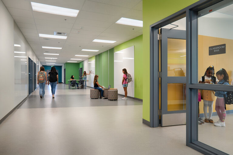 Students in a bright school hallway with whiteboard walls; two girls stand inside a classroom labeled B105.