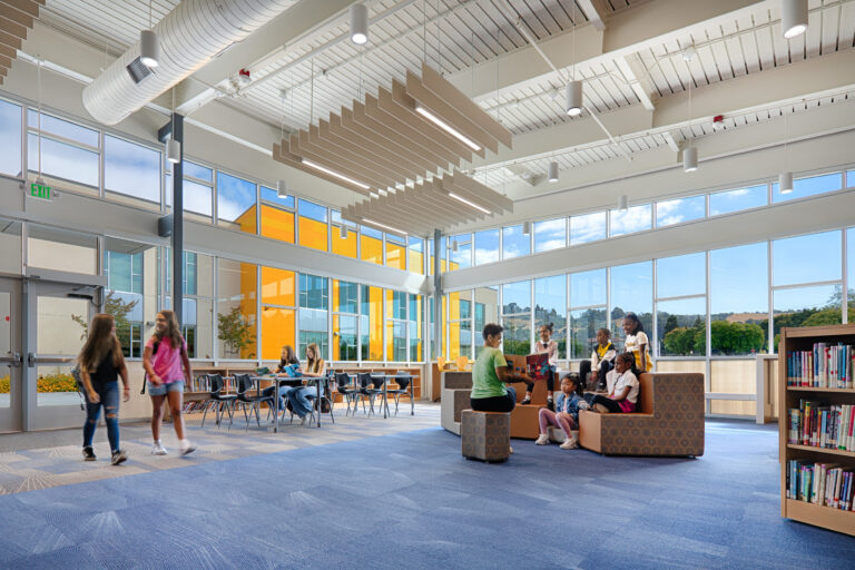 Children reading and walking in a bright modern school library with tall windows, tables, bookshelves, and lounge seating.