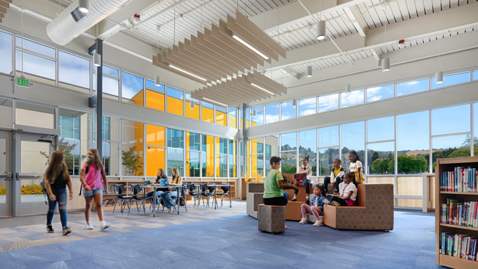 Children reading and walking in a bright modern school library with tall windows, tables, bookshelves, and lounge seating.