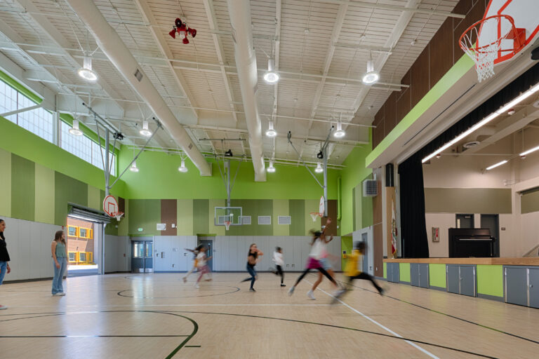 Children playing basketball in a bright school gym with green walls, multiple hoops, and a stage at one end.