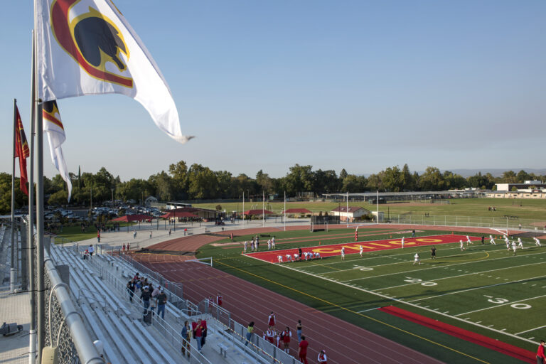 Wide view of a high school football stadium with players warming up, a running track, bleachers, and flags overhead
