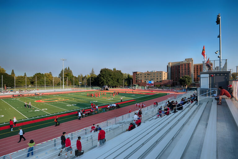 Wide view of a football stadium with players on the field, a track, and mostly empty bleachers under a clear blue sky