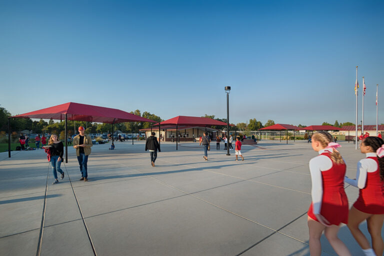 People walk across a large plaza with red shade canopies, a small building, and cheerleaders in red uniforms.