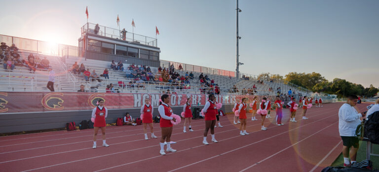 Cheerleaders lined up on a track in front of bleachers and a press box at a school stadium during sunset.