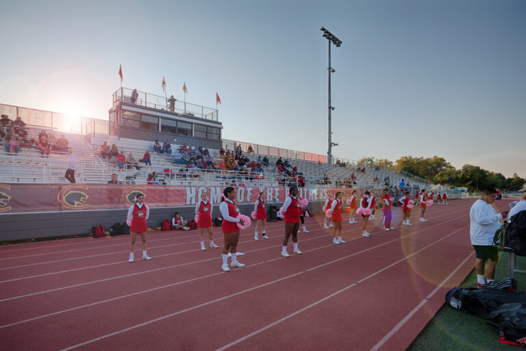 Cheerleaders lined up on a track in front of bleachers and a press box at a school stadium during sunset.