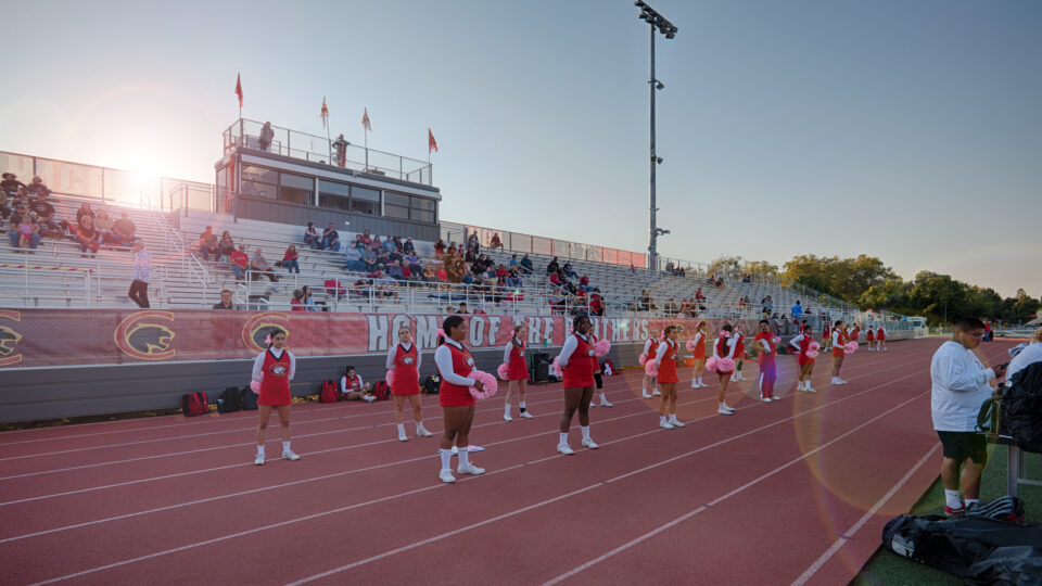 Cheerleaders lined up on a track in front of bleachers and a press box at a school stadium during sunset.