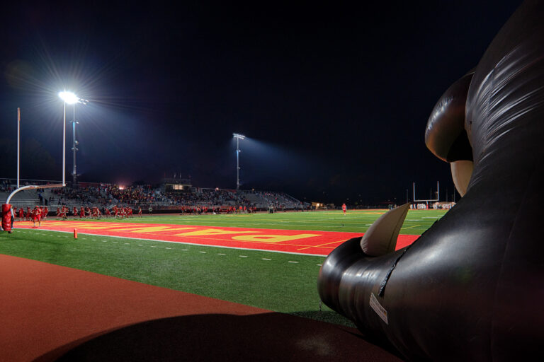 Night football field with players, bleachers, and stadium lights, framed by a large inflatable tunnel at the end zone.