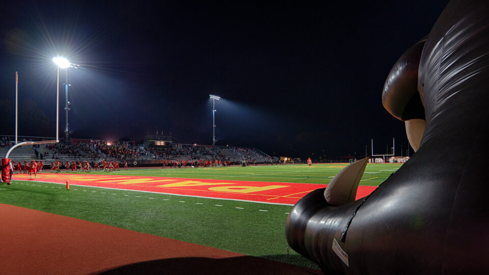 Night football field with players, bleachers, and stadium lights, framed by a large inflatable tunnel at the end zone.
