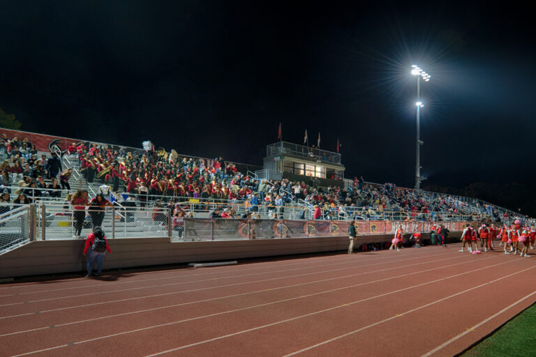 Crowd in stadium bleachers at night with a marching band, bright field lights, and cheerleaders by the track