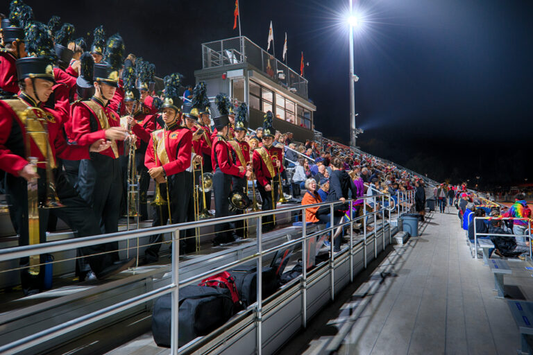 Marching band in red uniforms standing in stadium bleachers at a nighttime football game with a crowd nearby