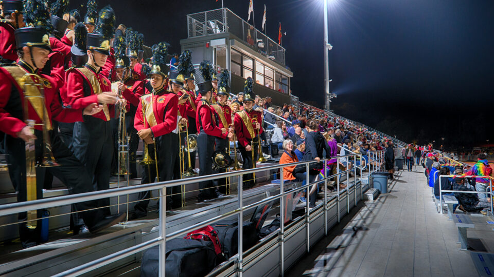 Marching band in red uniforms standing in stadium bleachers at a nighttime football game with a crowd nearby