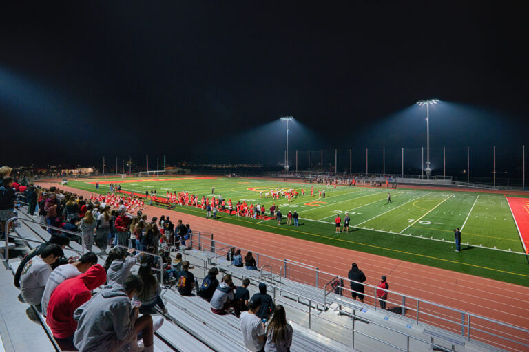 Nighttime football game on a brightly lit field with players, fans in the bleachers, and a track around the stadium