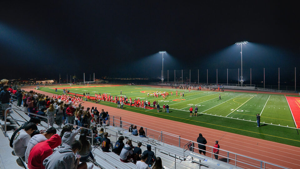 Nighttime football game on a brightly lit field with players, fans in the bleachers, and a track around the stadium