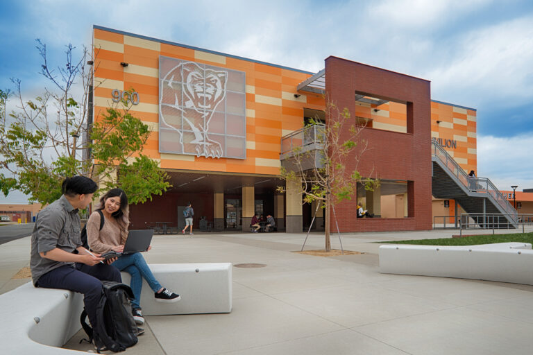 Two students with a laptop sit outside a modern school building with a large bear mural and orange-striped facade.
