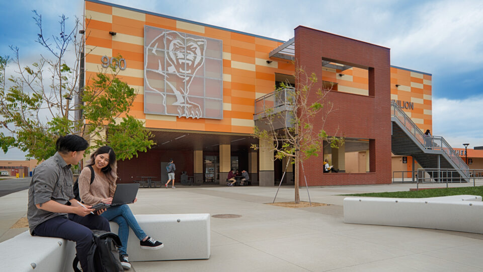 Two students with a laptop sit outside a modern school building with a large bear mural and orange-striped facade.
