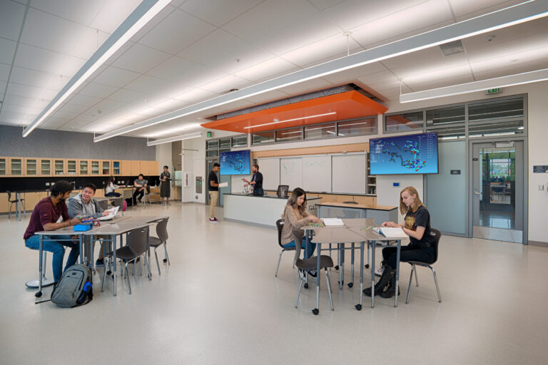 Students working and talking in a modern science classroom with lab tables, cabinets, and molecular models on screens