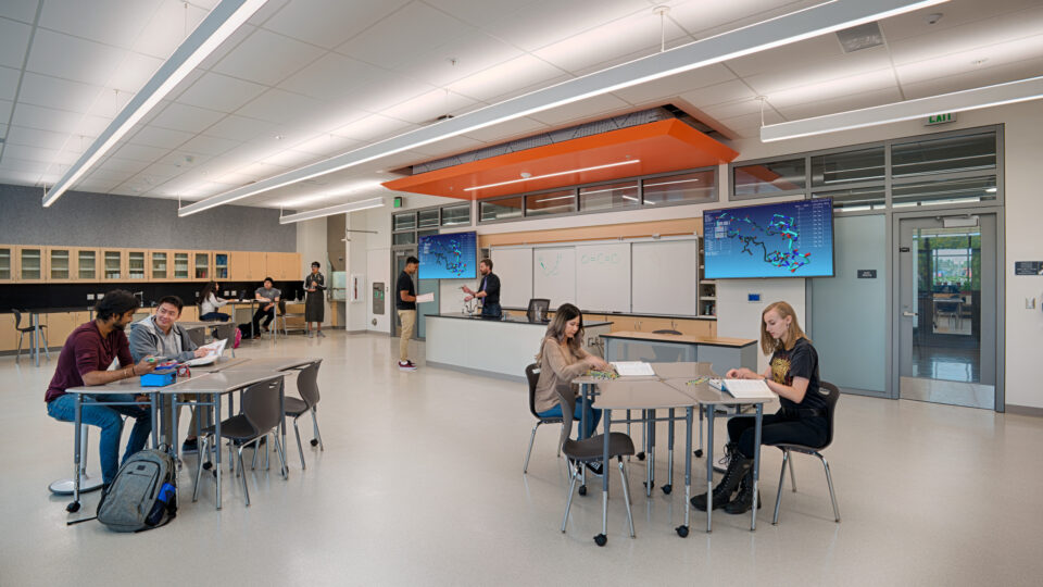 Students working and talking in a modern science classroom with lab tables, cabinets, and molecular models on screens