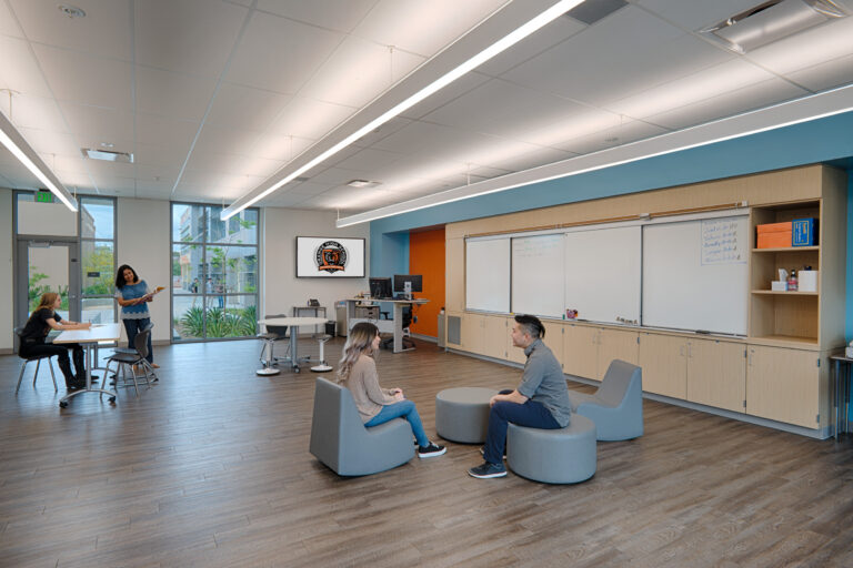 Modern high school classroom with flexible seating, whiteboards, and students talking with a teacher near large windows