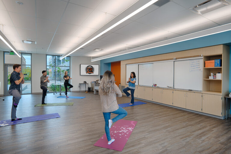 Five people practice tree pose on yoga mats in a bright classroom with large windows and whiteboards.