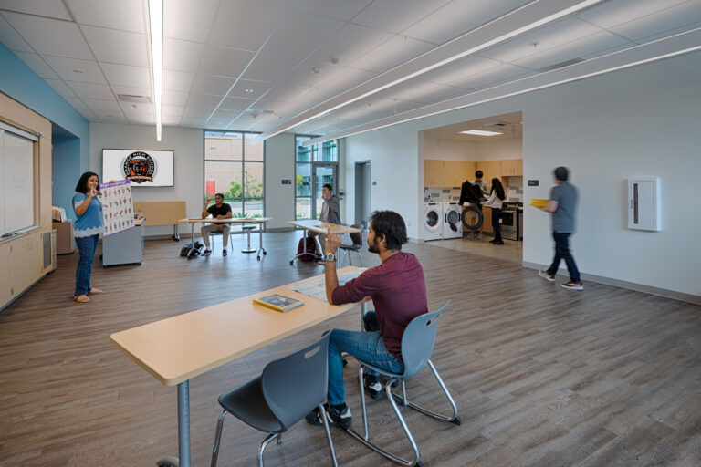 Teacher shows a sign language chart to students in a modern classroom with tables, large windows, and a kitchenette.