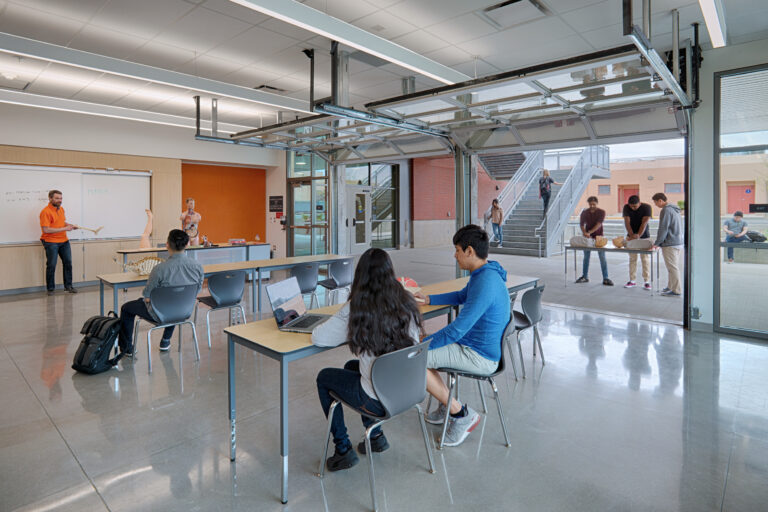 Anatomy class in a modern lab with students seated indoors and others practicing CPR outside through an open glass wall