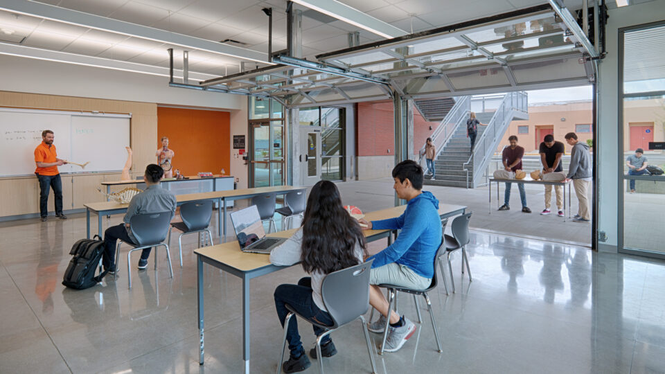 Anatomy class in a modern lab with students seated indoors and others practicing CPR outside through an open glass wall