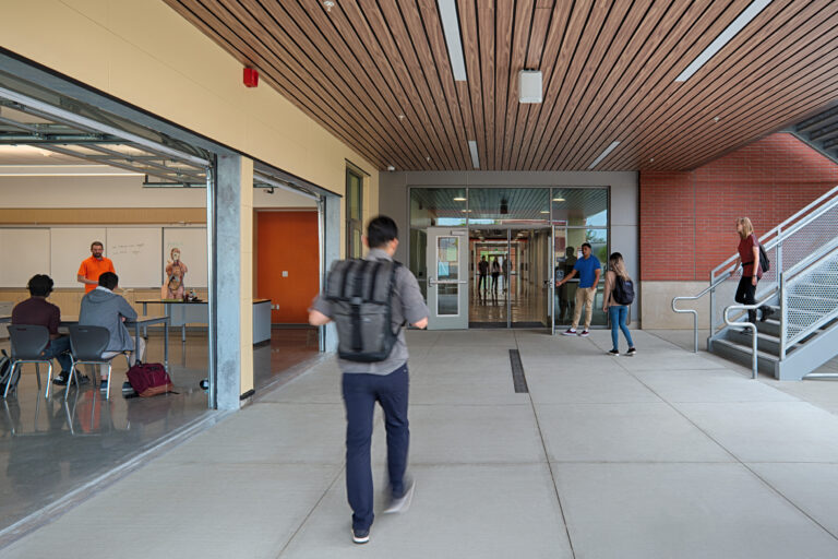Students walking through a modern campus breezeway beside an open classroom where an anatomy lesson is in progress