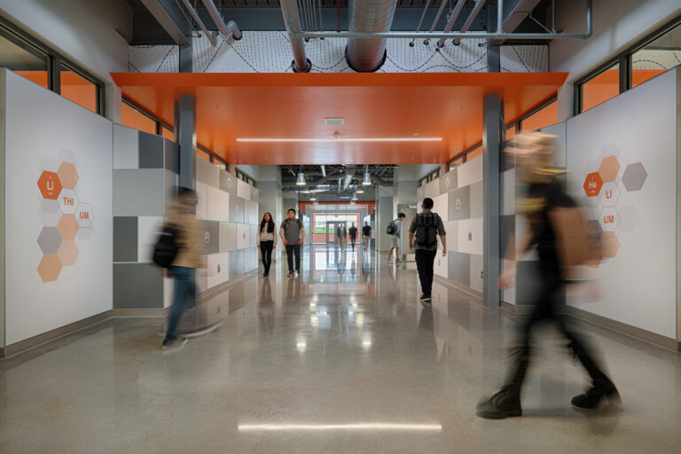 Students walking through a modern hallway with orange ceiling panels and hexagonal chemistry graphics on the walls