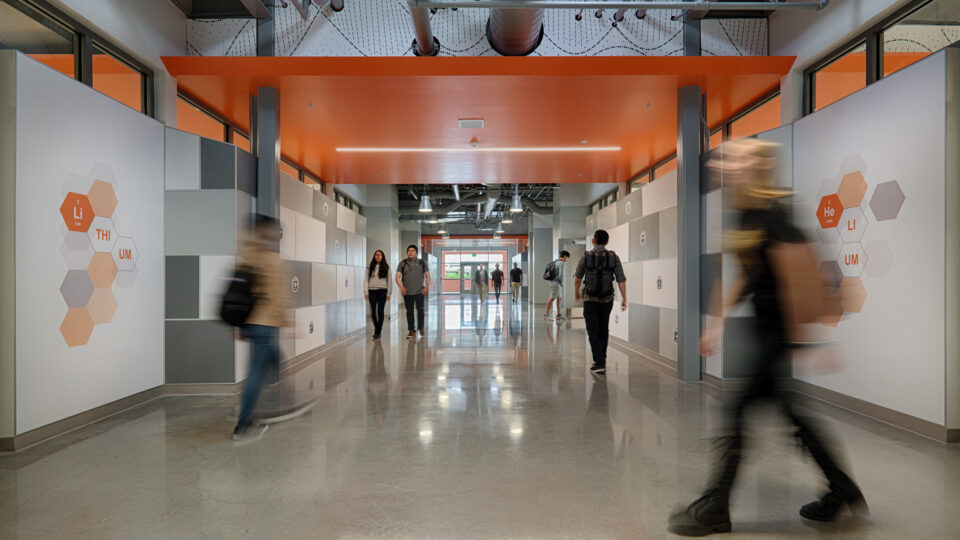 Students walking through a modern hallway with orange ceiling panels and hexagonal chemistry graphics on the walls