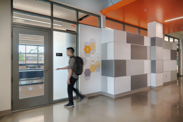 Student walking past a chemistry lab in a modern school hallway with geometric wall panels and an orange ceiling