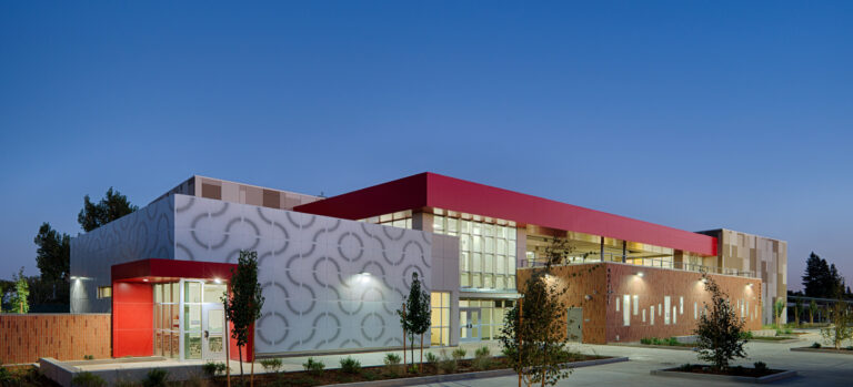 Modern school building at dusk with red accents, patterned white facade, large windows, and a landscaped courtyard