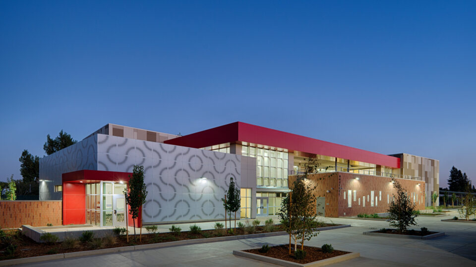 Modern school building at dusk with red accents, patterned white facade, large windows, and a landscaped courtyard