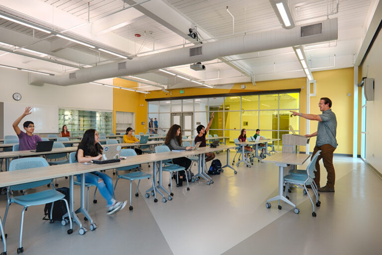 Teacher leading a classroom discussion as several students raise their hands in a bright modern room with laptops.