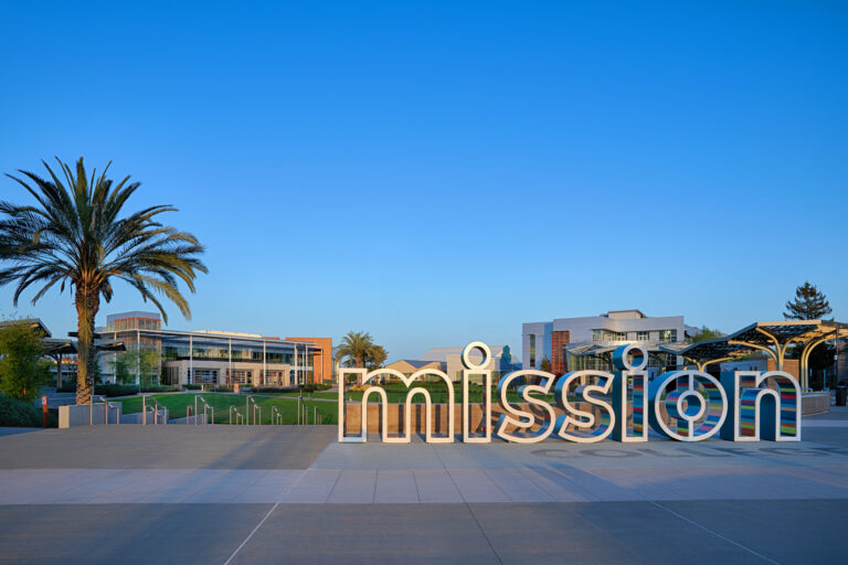 Large colorful Mission sign in front of a modern campus plaza with palm trees and buildings under a clear blue sky