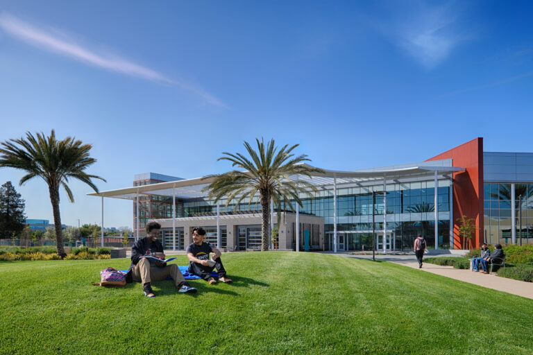 Two students sitting on a campus lawn in front of a modern glass building and palm trees under a clear blue sky